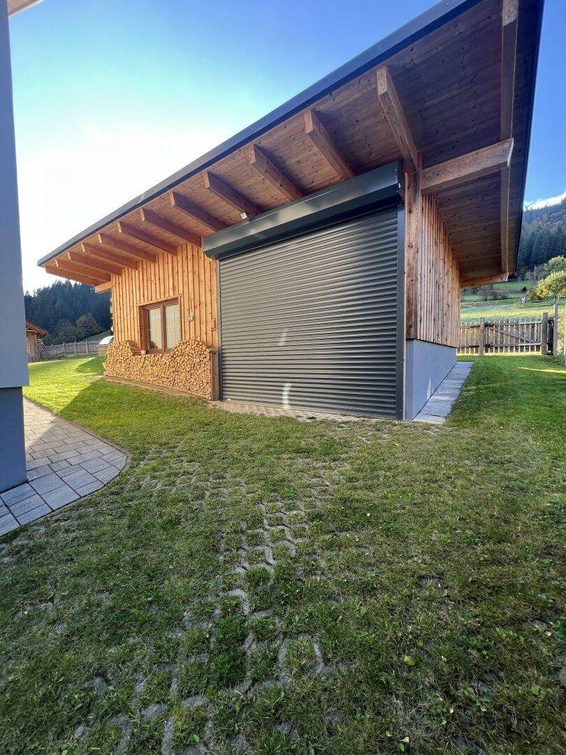 Seitenansicht eines Holzgebäudes mit großem Rolltor und gestapeltem Brennholz auf grünem Rasen.
