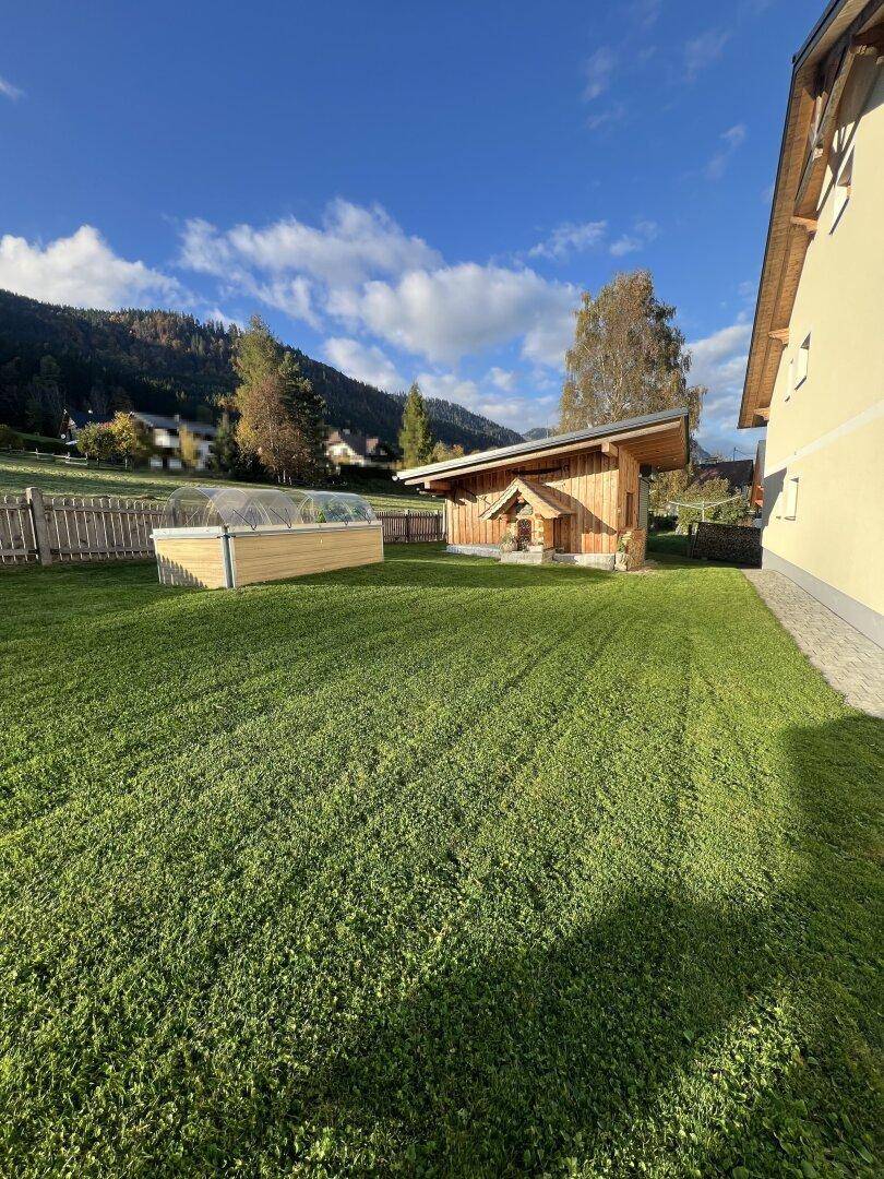 Gepflegter Gartenbereich mit Rasenfläche, einem Holzschuppen und einem erhöhten Pflanzenbeet vor Bergkulisse.