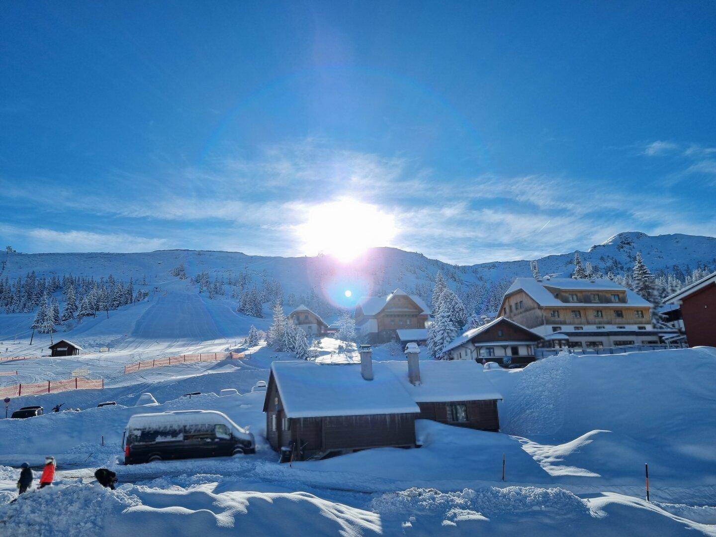Panoramablick auf ein verschneites Bergdorf mit Skigebiet und strahlendem Sonnenschein im Winter.