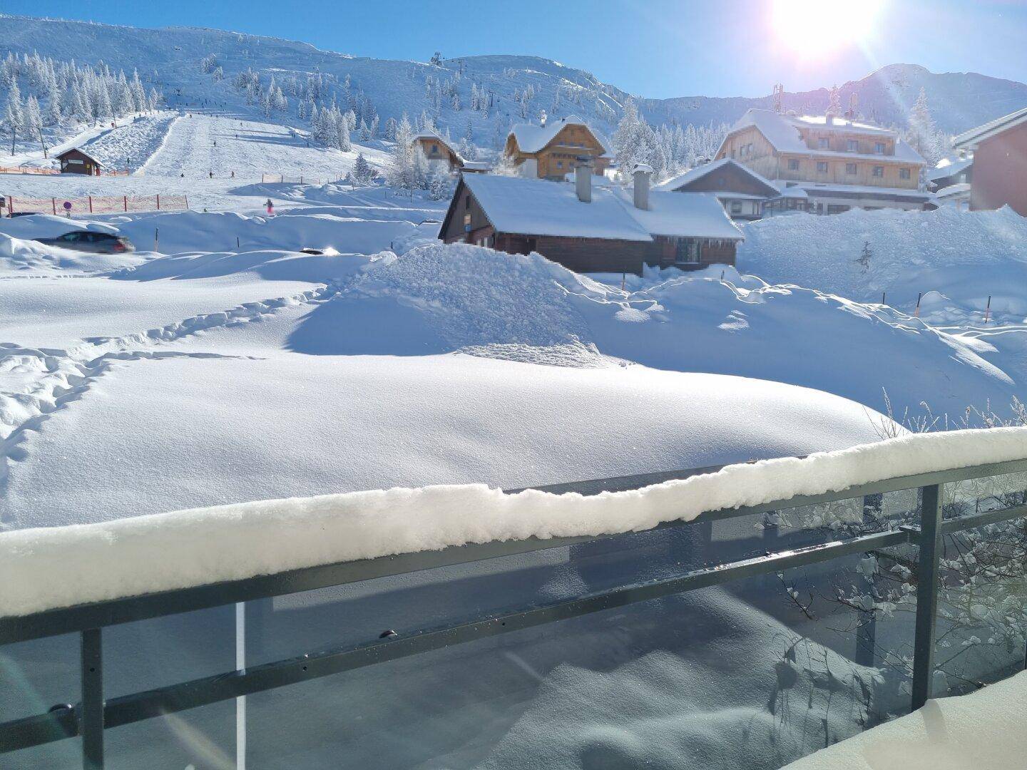 Blick von einem schneebedeckten Balkon auf eine winterliche Berglandschaft mit Skihängen und Häusern.