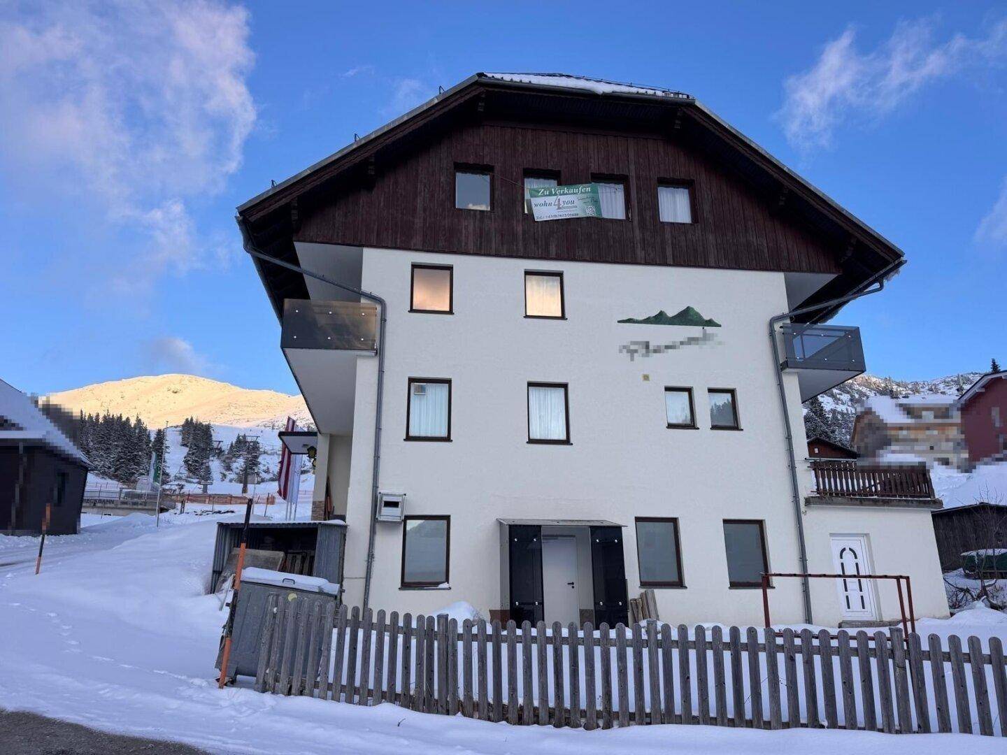 Weißes Gebäude mit Holzelementen und Balkonen in einer verschneiten Berglandschaft, umgeben von einem Zaun.