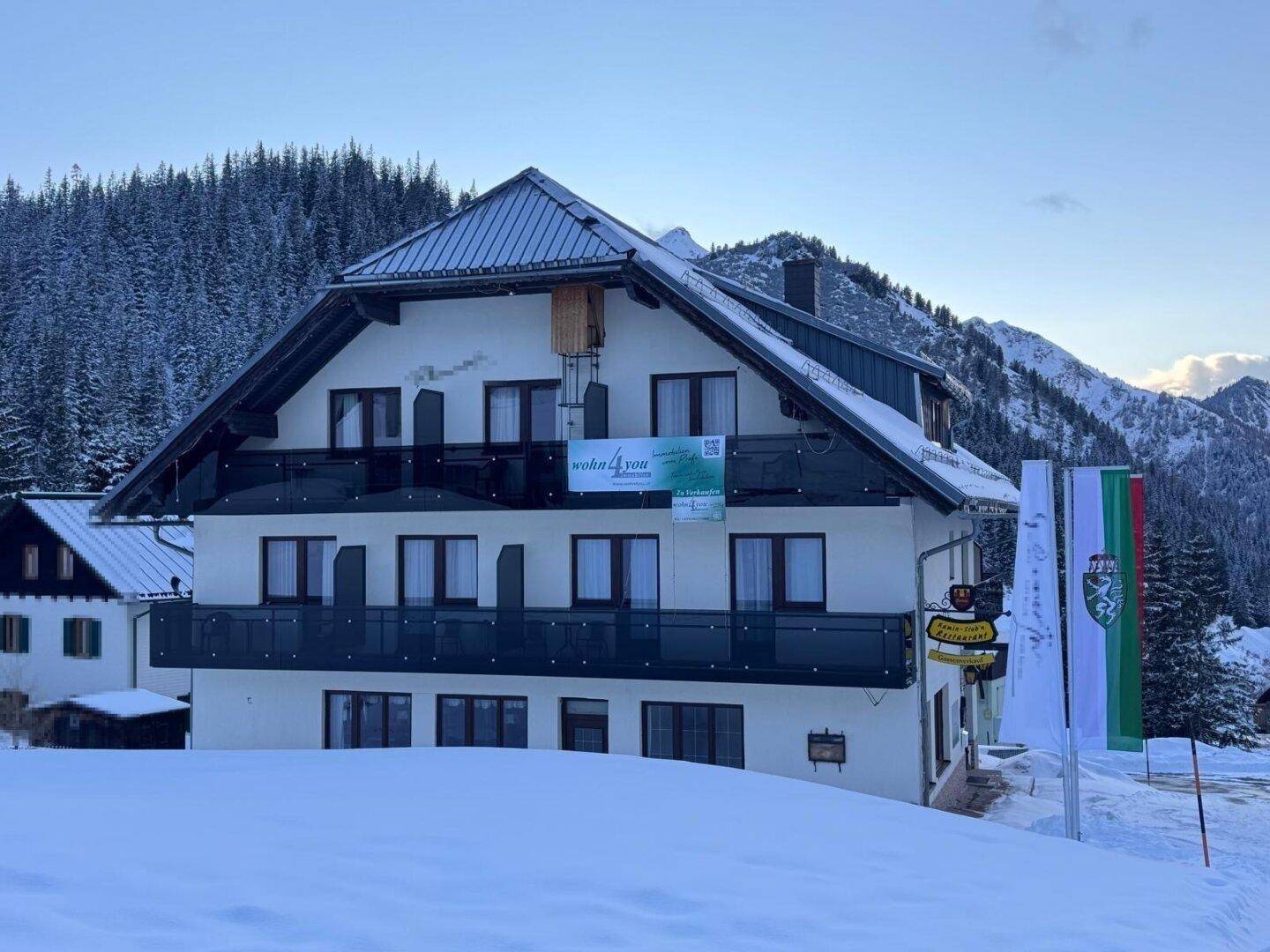 Mehrfamilienhaus in winterlicher Bergkulisse mit Balkonen und traditioneller Architektur, umgeben von Schnee.