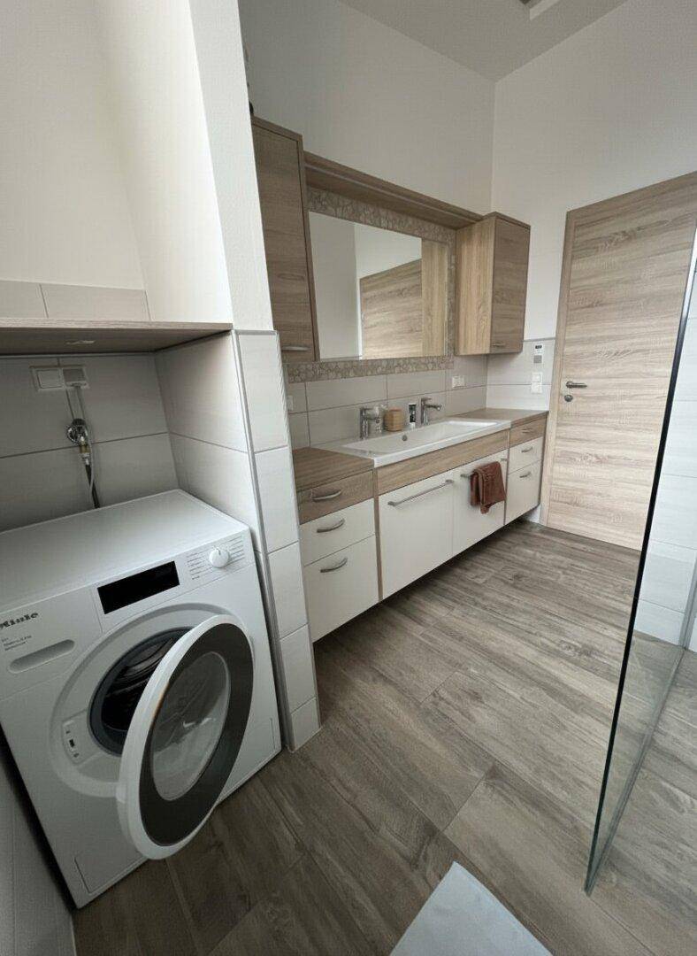 Badezimmer mit Einzelwaschbecken, Spiegelschrank und integrierter Waschmaschine auf Holzoptik-Boden.