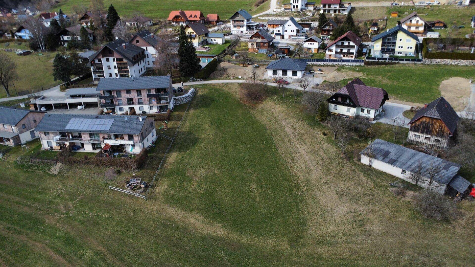 Weitläufige Luftaufnahme eines Dorfes mit vielen Wohnhäusern, eingebettet in eine grüne Hügellandschaft.