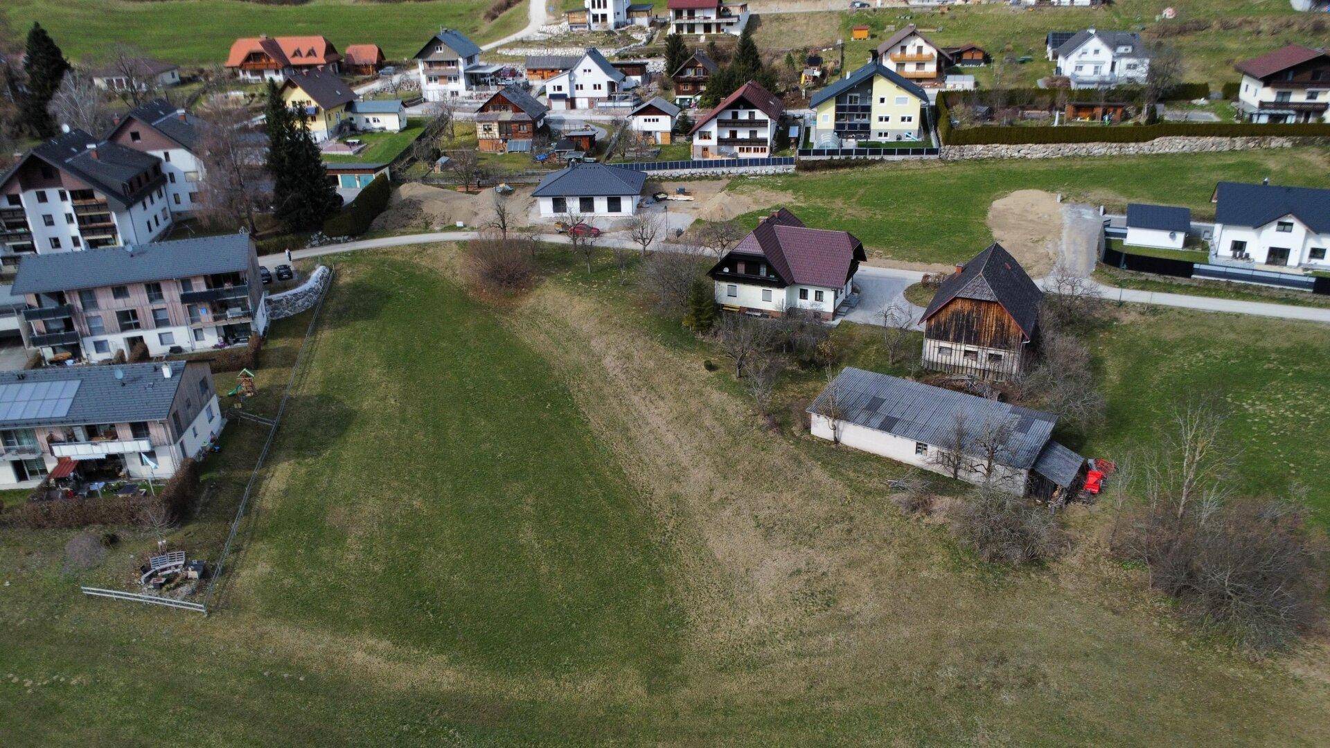 Weitläufige Luftaufnahme eines Dorfes mit vielen Wohnhäusern, eingebettet in eine grüne Hügellandschaft.