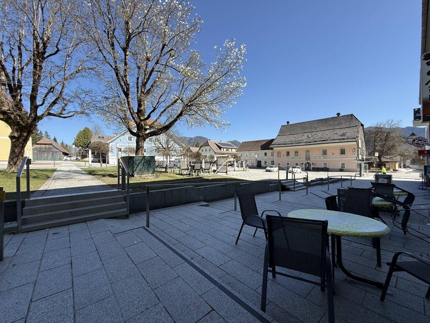 Gepflasterter Platz mit Tischen und Stühlen, umgeben von historischen Gebäuden und Bäumen.