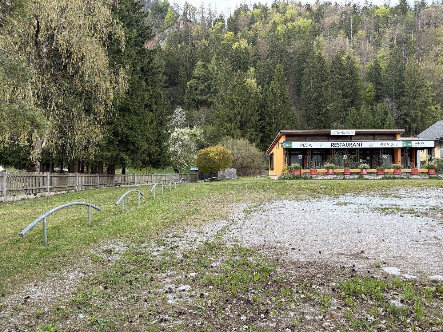 Gebäude mit Restaurantfassade, umgeben von einer Schotterfläche und Rasen, mit Wald im Hintergrund.