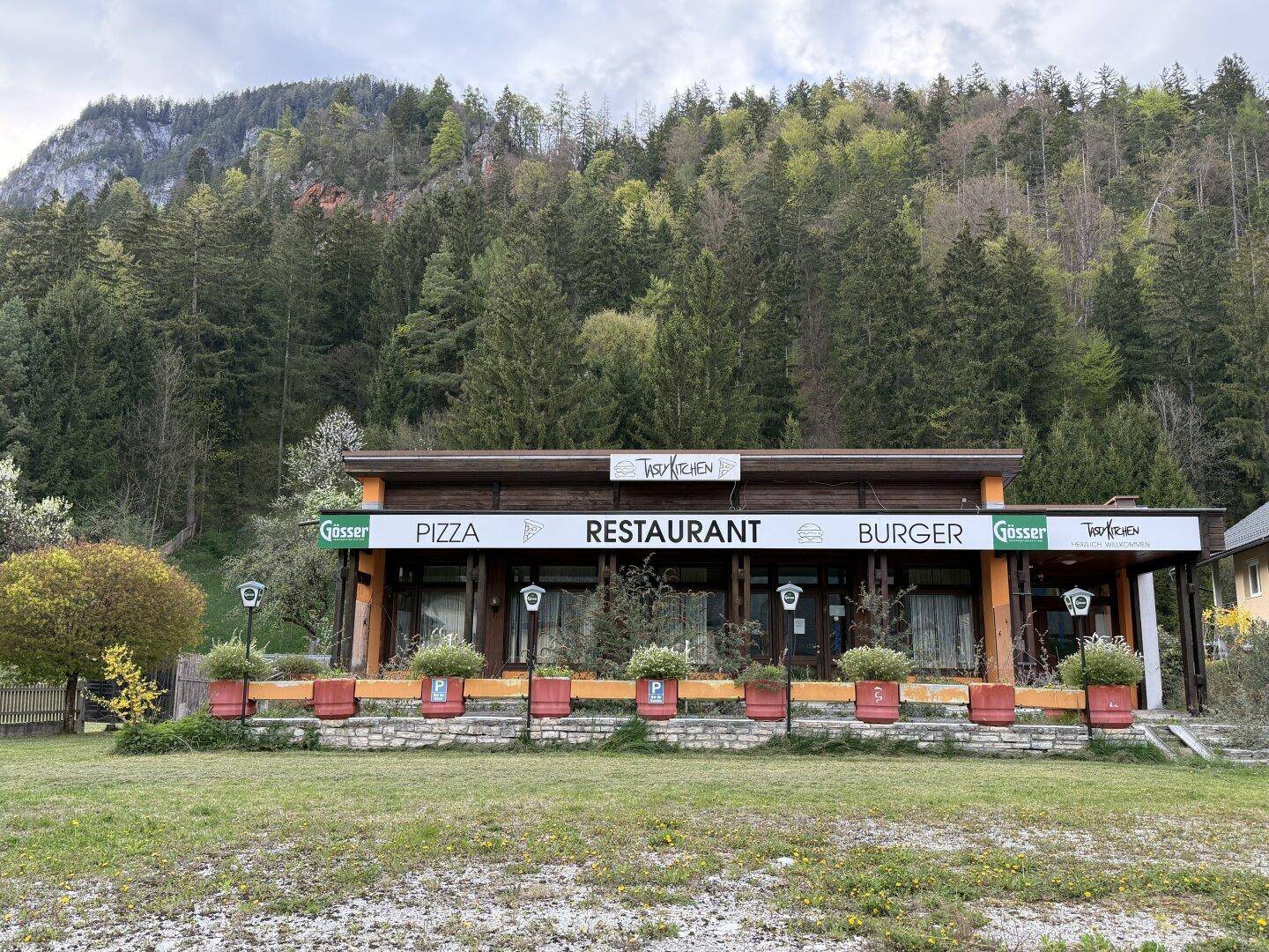 Vorderansicht des Restaurants mit großen Fenstern, Topfpflanzen und einem Parkplatzbereich.