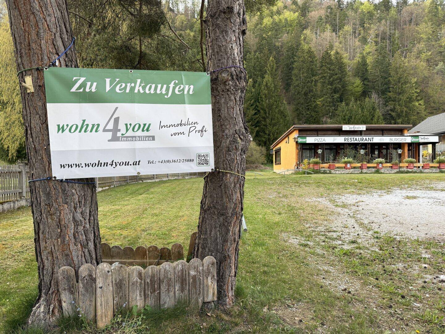 Verkaufsschild an Bäumen vor einem Gebäude mit Wald im Hintergrund, umgeben von Grünflächen.