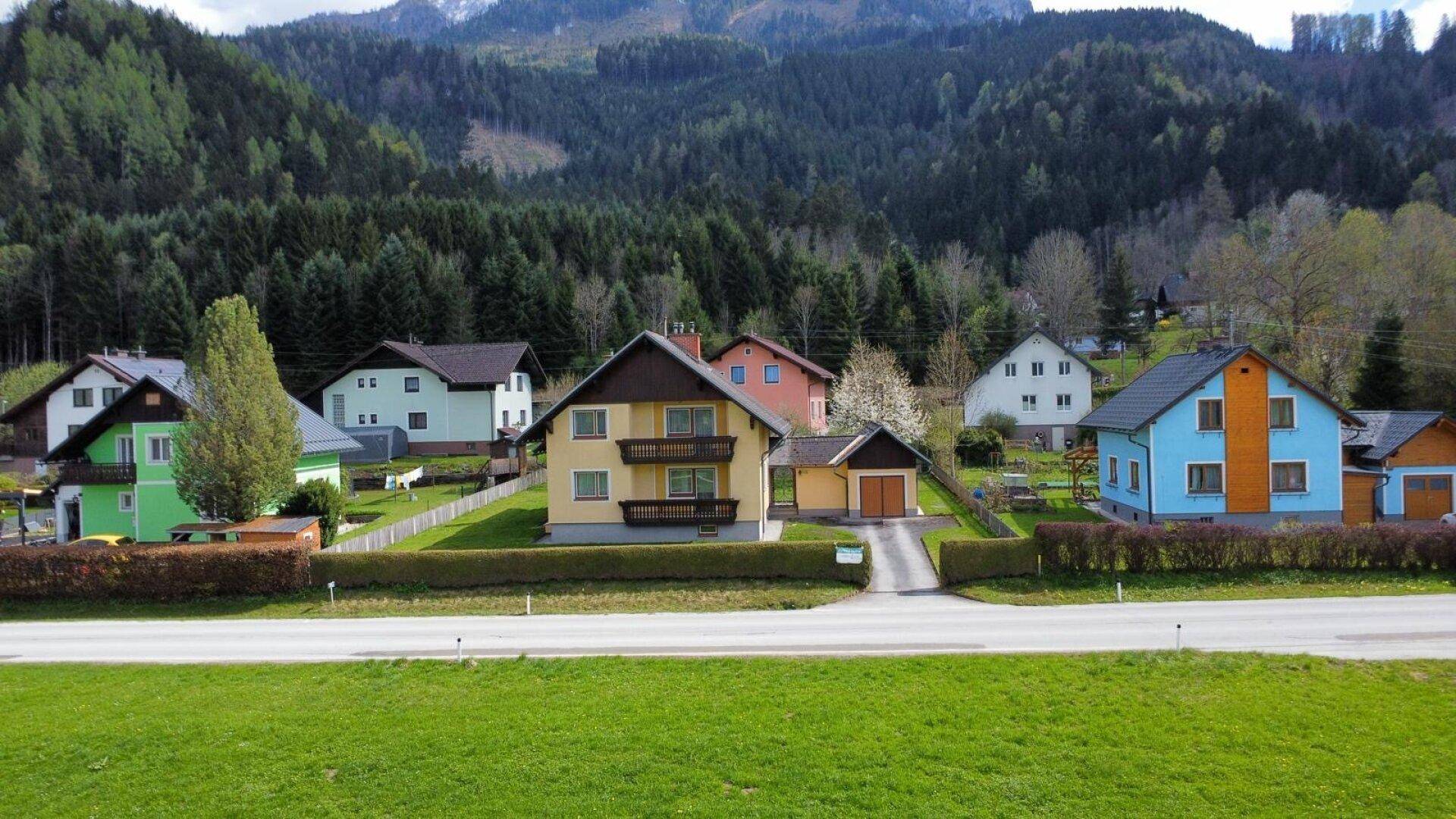 Luftaufnahme einer ländlichen Siedlung mit verschiedenen Häusern vor bewaldeter Bergkulisse.