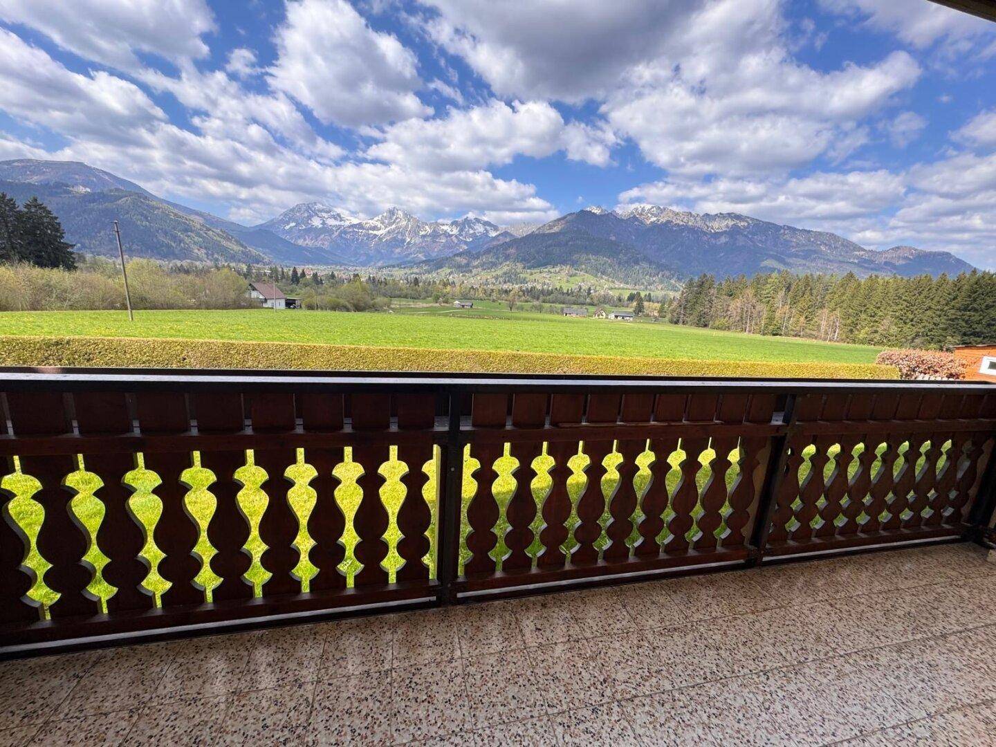 Blick vom Balkon über grüne Felder auf die beeindruckende Bergkulisse unter blauem Himmel.