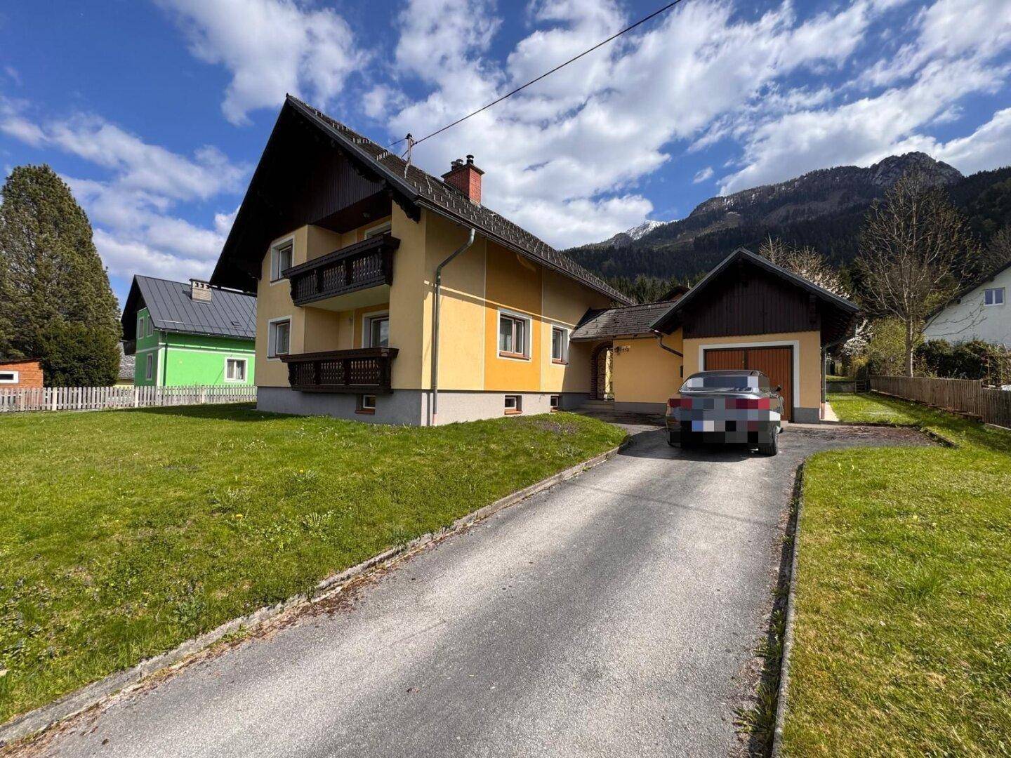 Einfamilienhaus mit Zufahrt und integrierter Garage, umgeben von grüner Landschaft und Bergen.