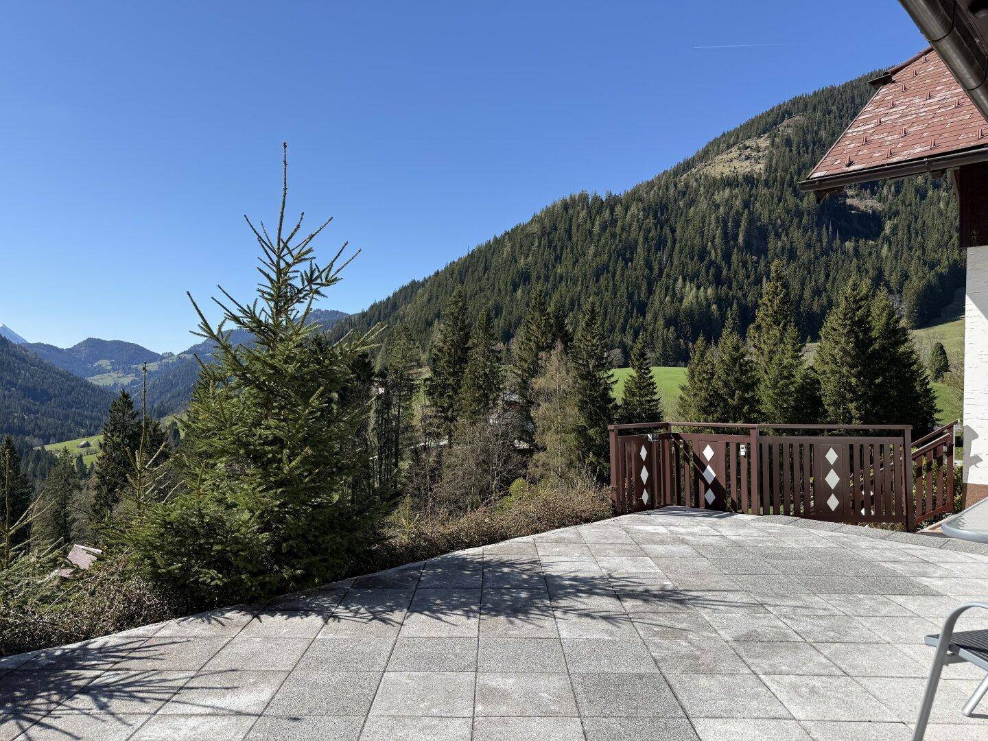 Große gepflasterte Terrasse mit Holzzaun und weitem Blick auf die bewaldete Berglandschaft.