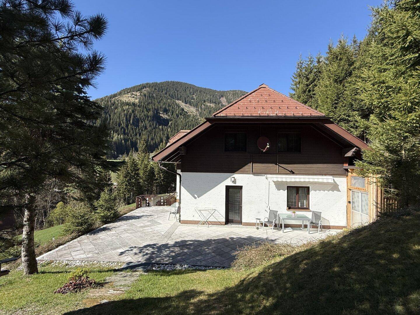 Charmantes Haus mit traditionellem Dach und großer Terrasse inmitten einer idyllischen Berglandschaft.