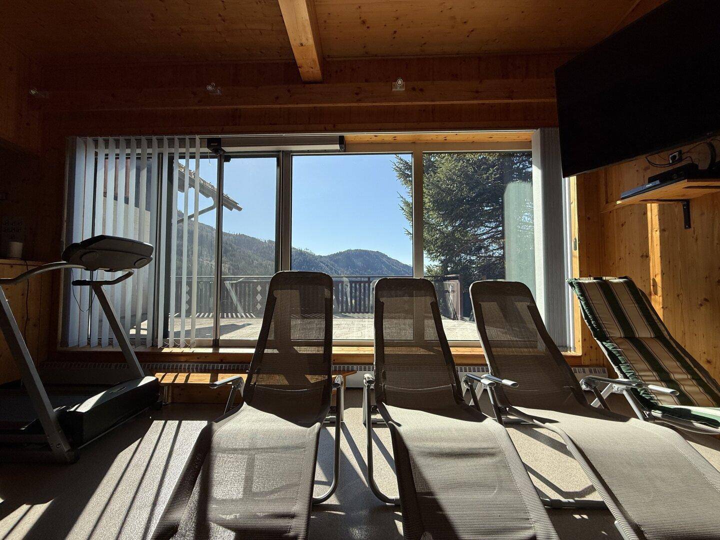 Heller Fitnessraum mit Holzvertäfelung, Liegestühlen und Zugang zu einer Terrasse mit Bergblick.