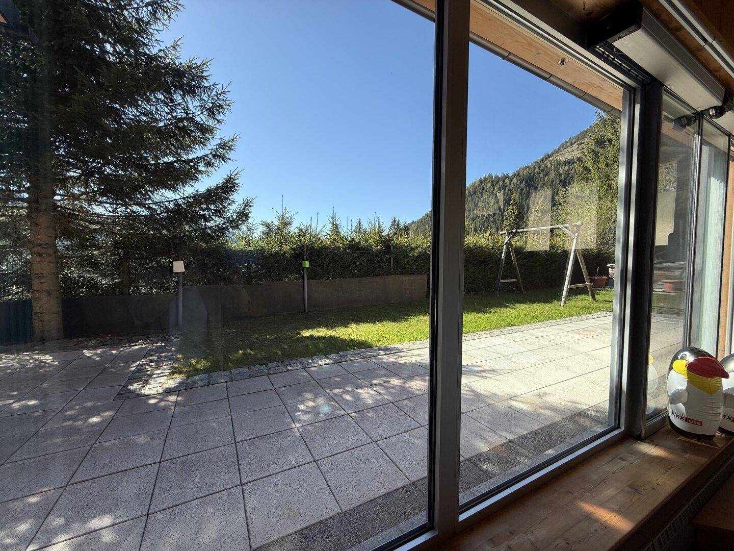 Große Fensterfront mit Blick auf den grünen Garten und eine Schaukel im Freien.