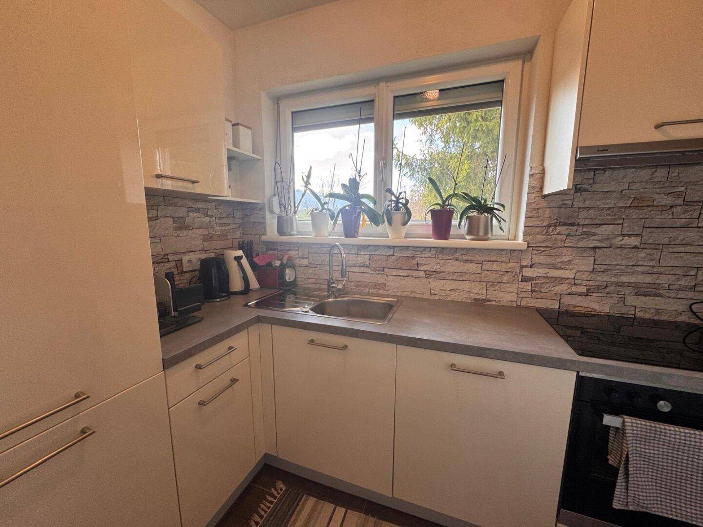 Moderne Küche mit weißen Hochglanzschränken, Steinoptik-Rückwand und Fensterblick ins Grüne.