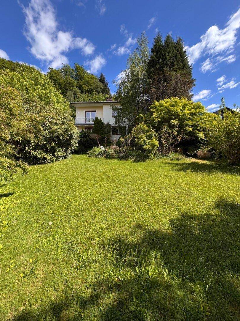 Sonniger Garten mit großer Rasenfläche und dem Wohnhaus, eingebettet in eine grüne Hügellandschaft.