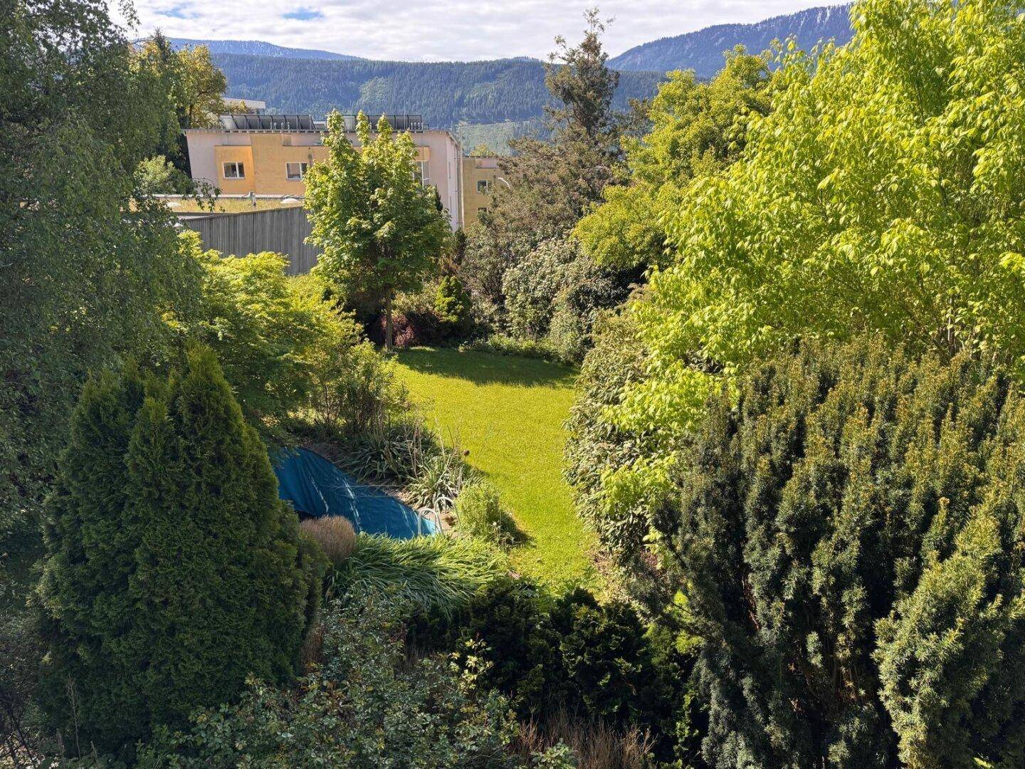 Blick von oben auf den gepflegten Garten mit Rasenfläche, umgeben von Bäumen und mit Bergsicht.