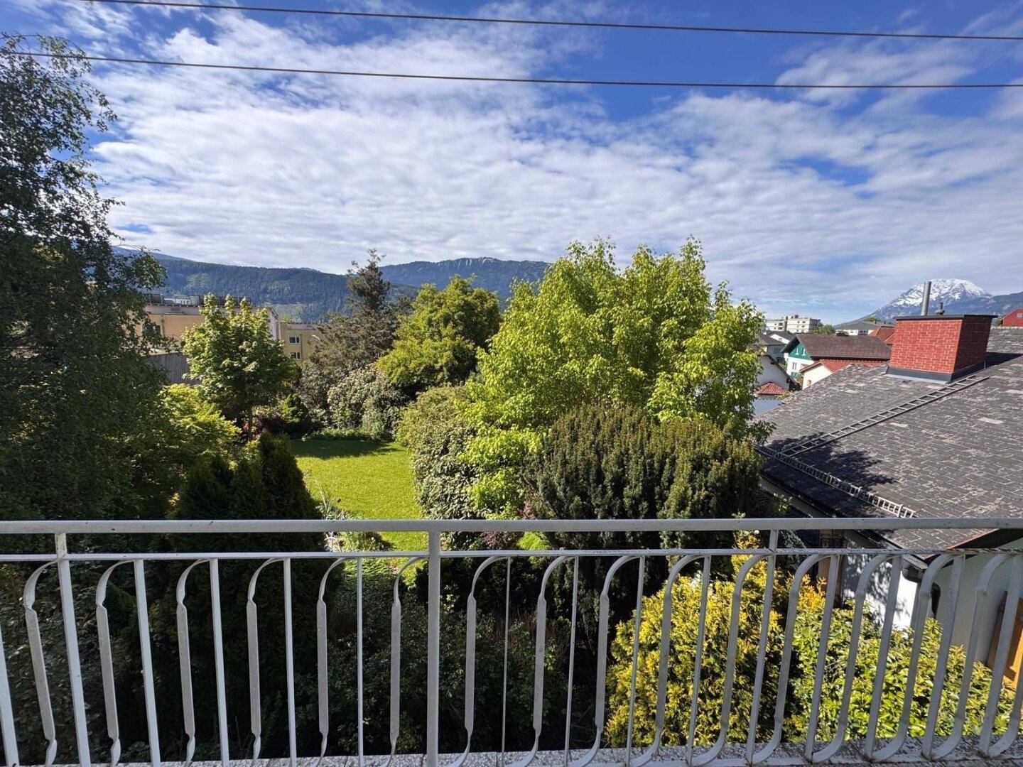 Panoramablick vom Balkon über den grünen Garten, die Dächer und die beeindruckende Berglandschaft.