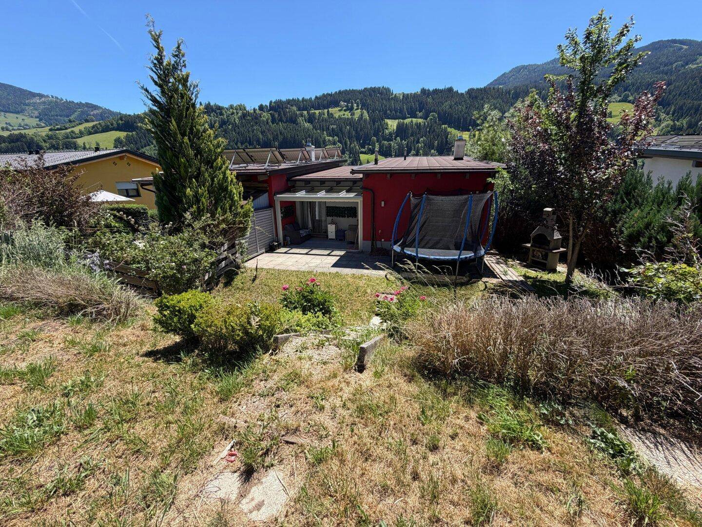 Großer Gartenbereich mit Trampolin und Grillplatz, umgeben von üppiger grüner Natur und Bergen.