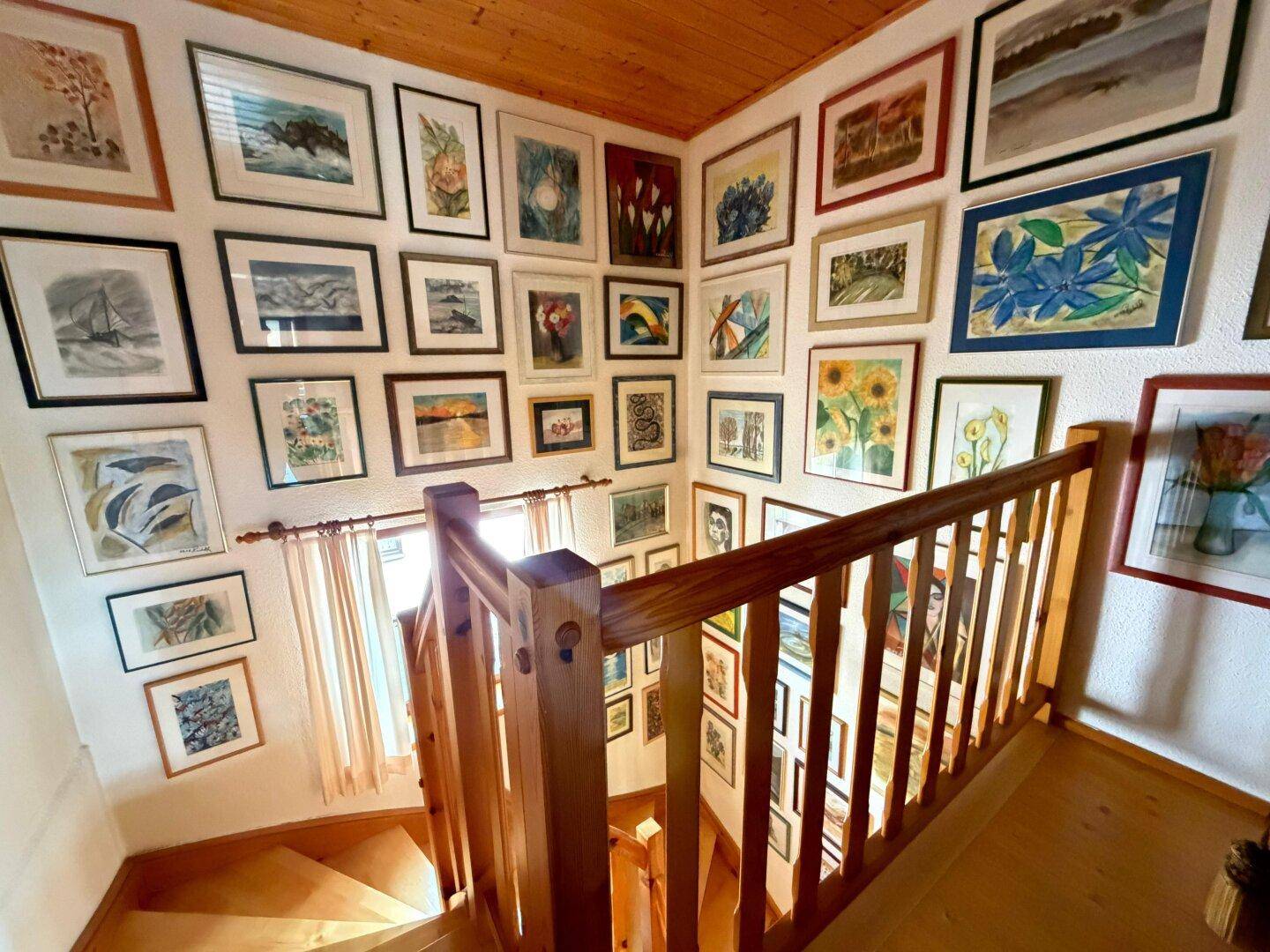 Heller Treppenaufgang mit Holzgeländer und einer Galerie von Kunstwerken an den Wänden.