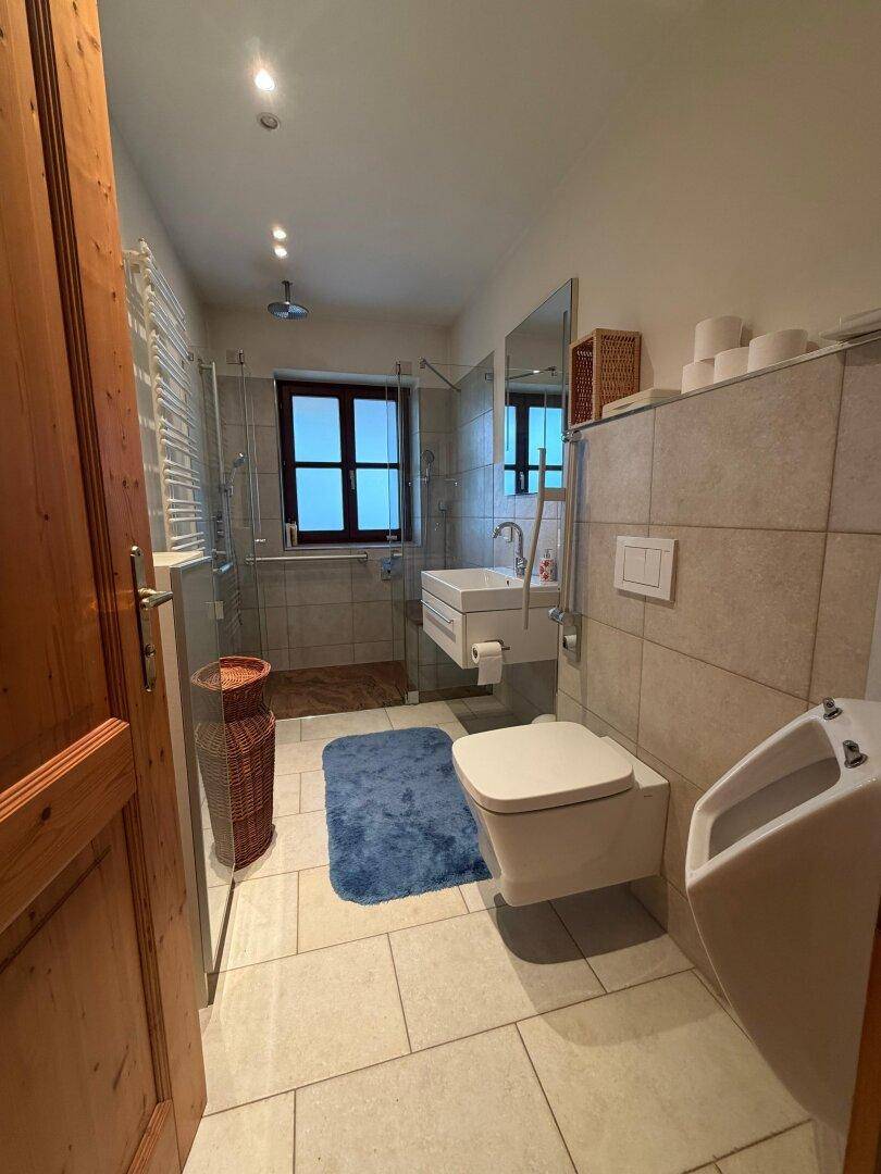 Modernisiertes Badezimmer mit Dusche, Wand-WC, Urinal und Fliesenboden.