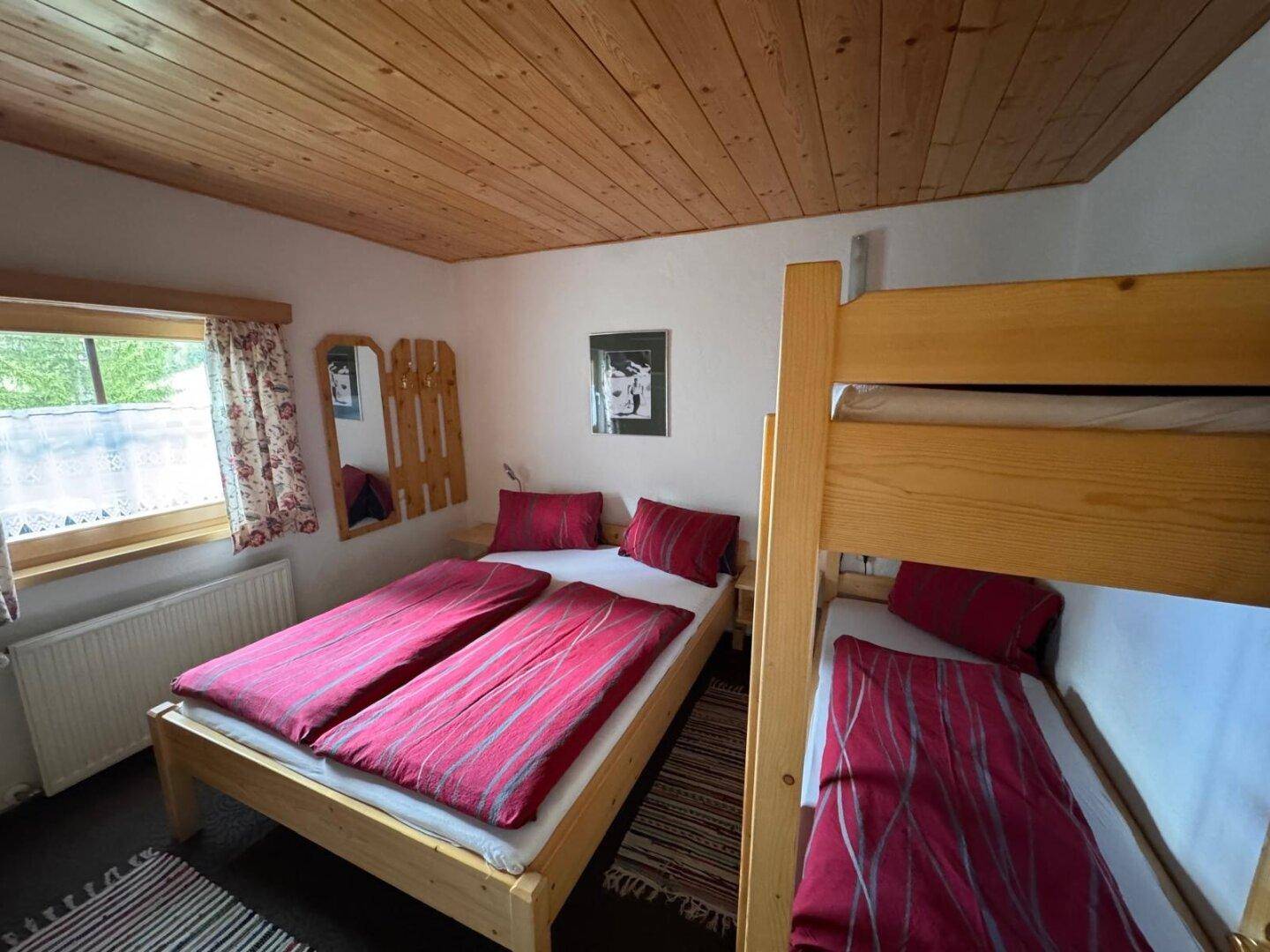 Schlafzimmer mit Doppelbett und Etagenbett, Holzbalkendecke und Fensterblick ins Grüne.