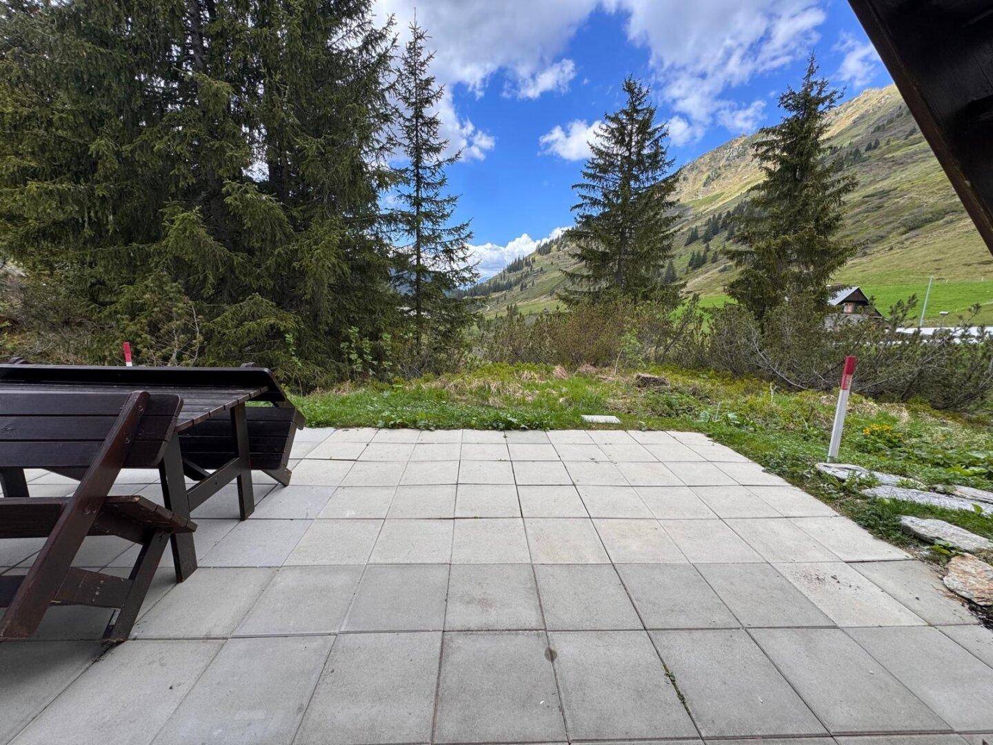 Gepflasterte Terrasse mit Holztisch und Bänken, umgeben von Bäumen und Bergblick.