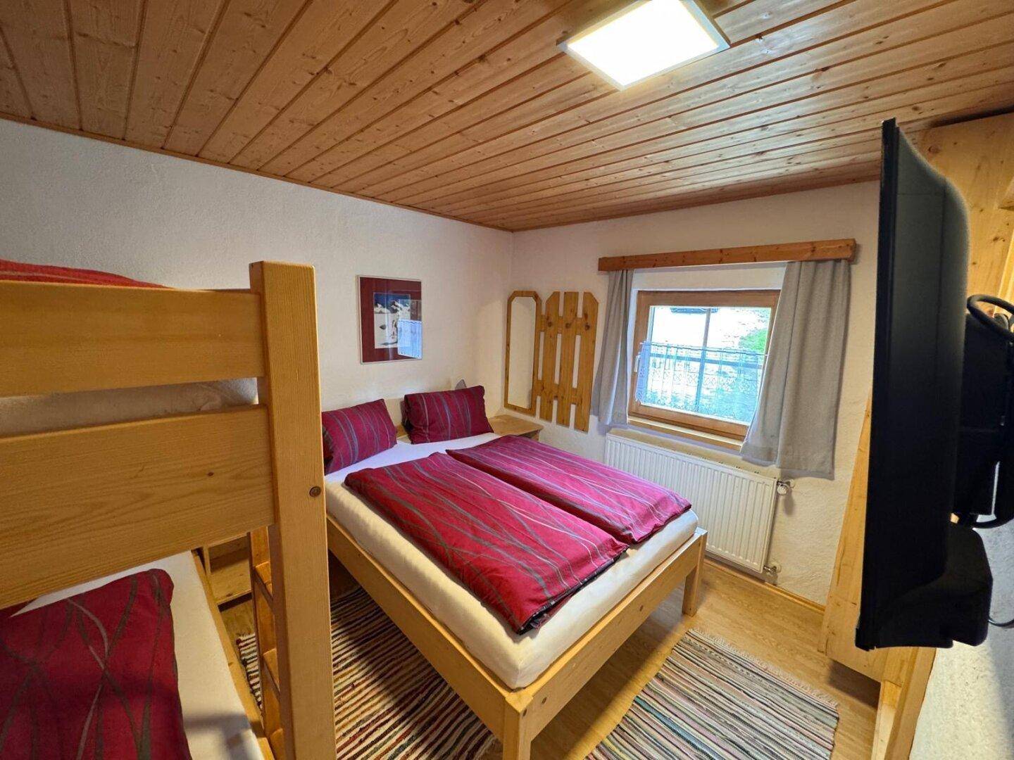 Schlafzimmer mit Doppelbett und Etagenbett, Holzbalkendecke und Fernseher an der Wand.
