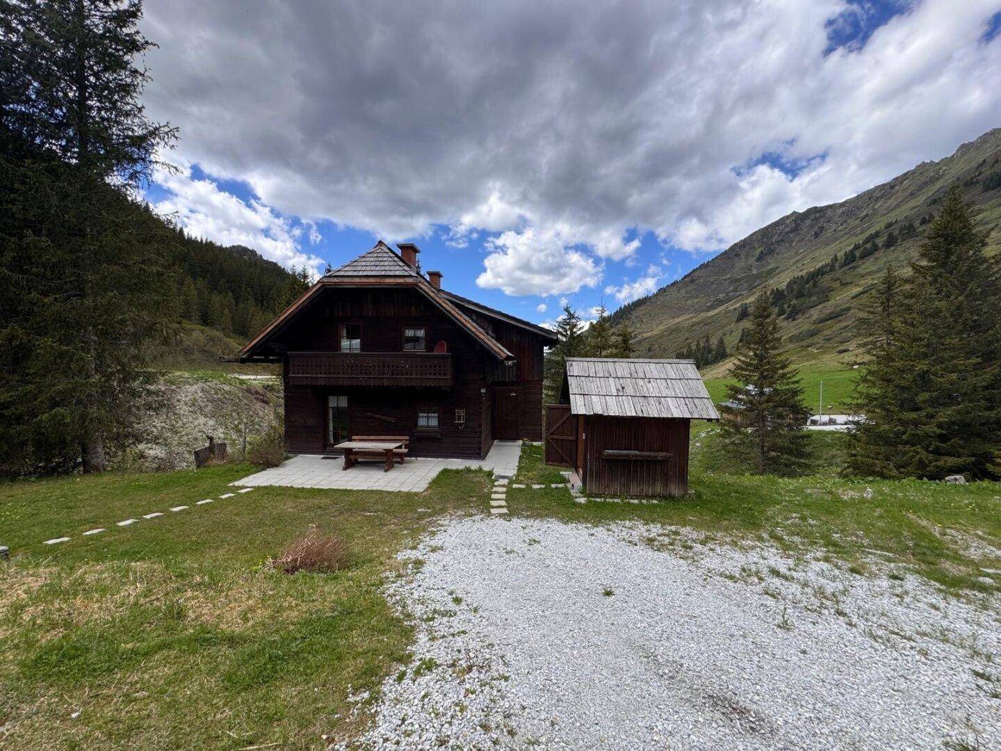 Alpines Holzhaus mit Schindeldach und kleiner Terrasse, umgeben von Bergen und Bäumen.