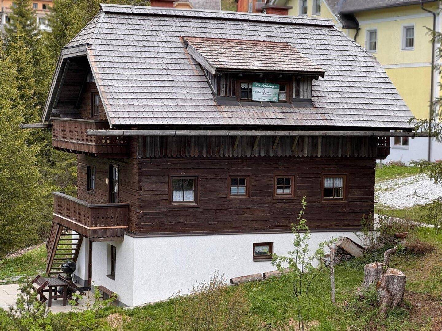 Traditionelles Holzhaus mit Schindeldach und Balkonen, teilweise weiß verputztem Sockel.
