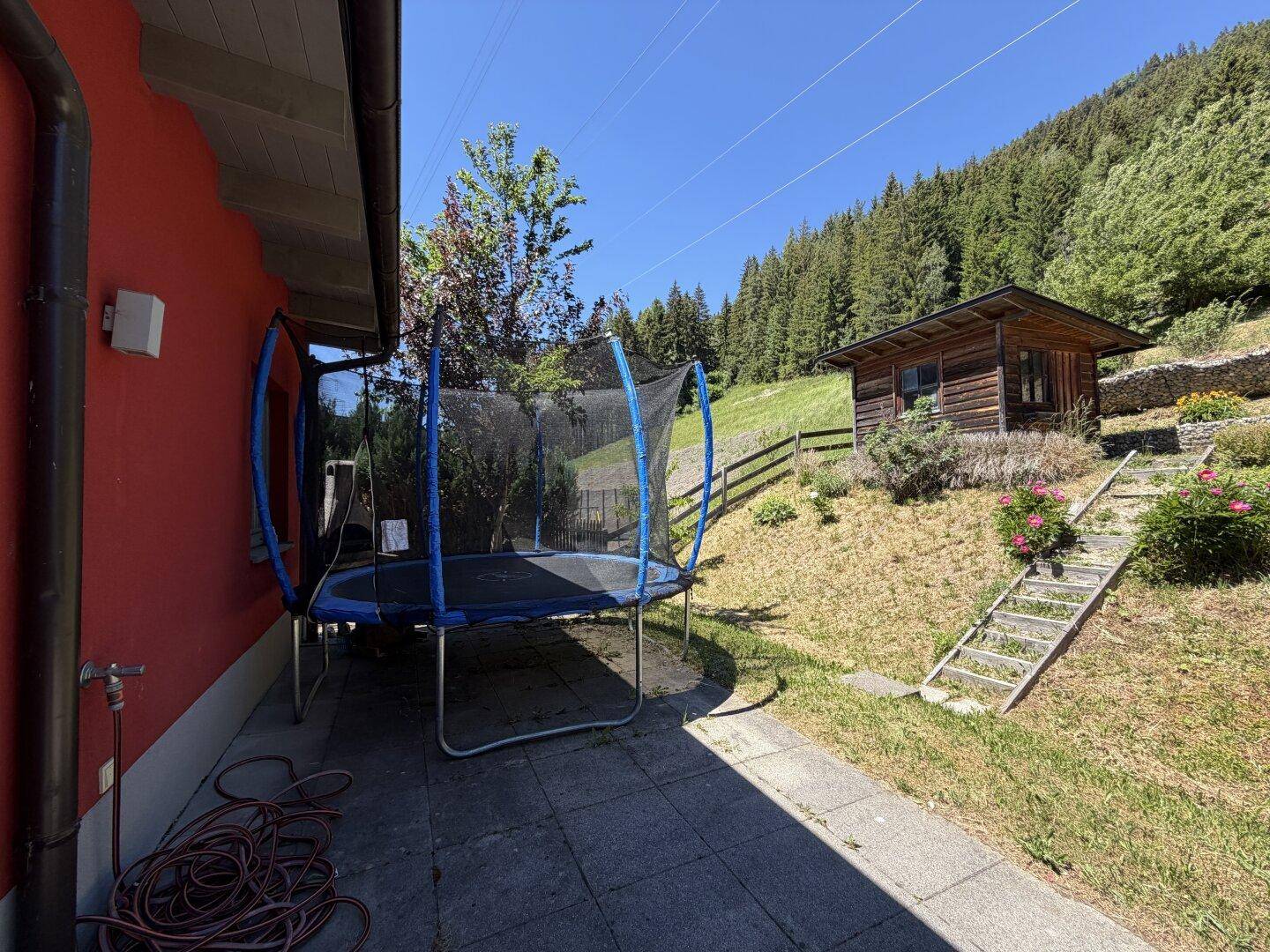 Gartenbereich mit Trampolin und kleinem Holzschuppen, umgeben von Natur und Bäumen.