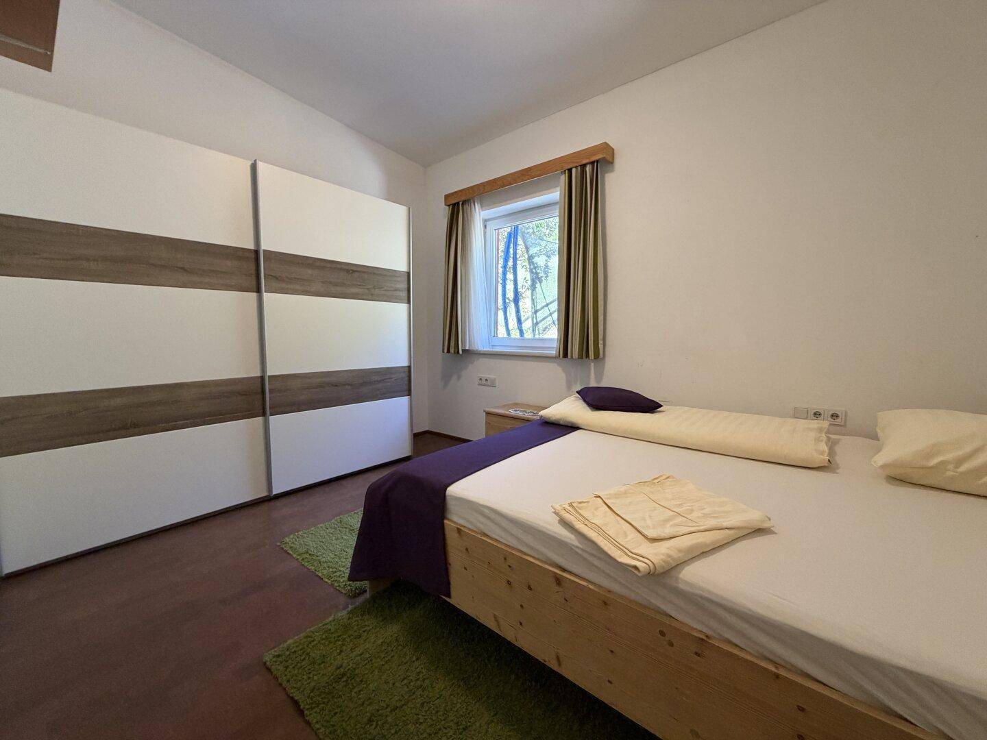 Helles Schlafzimmer mit Holzbett, Schiebetürenschrank und Blick ins Grüne.