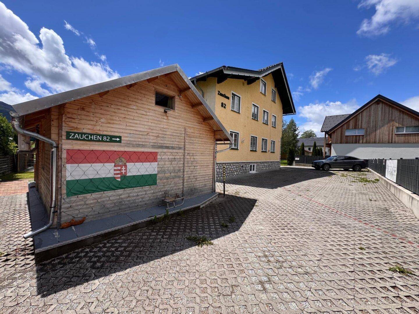 Holzschuppen mit ungarischer Flagge und großem gepflastertem Parkplatz neben dem Hauptgebäude.