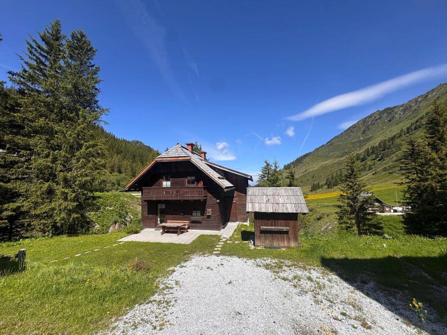Idyllisches Holzchalet umgeben von grünen Wiesen und Bergen unter strahlend blauem Himmel.
