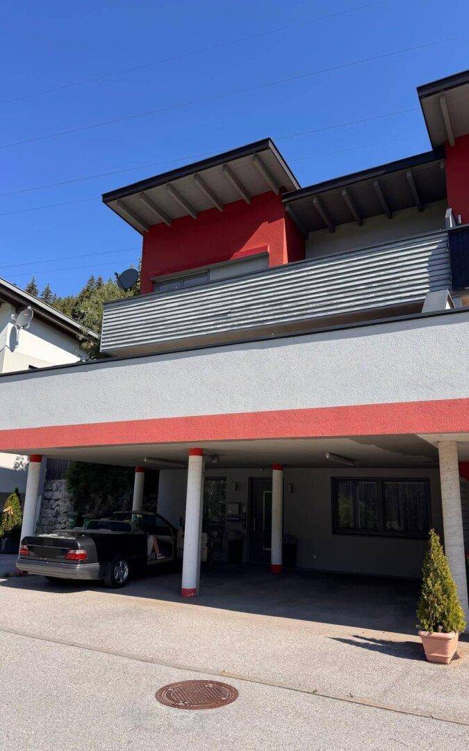 Modernes Gebäude mit roter und grauer Fassade, überdachtem Carport und Balkon im oberen Stockwerk.