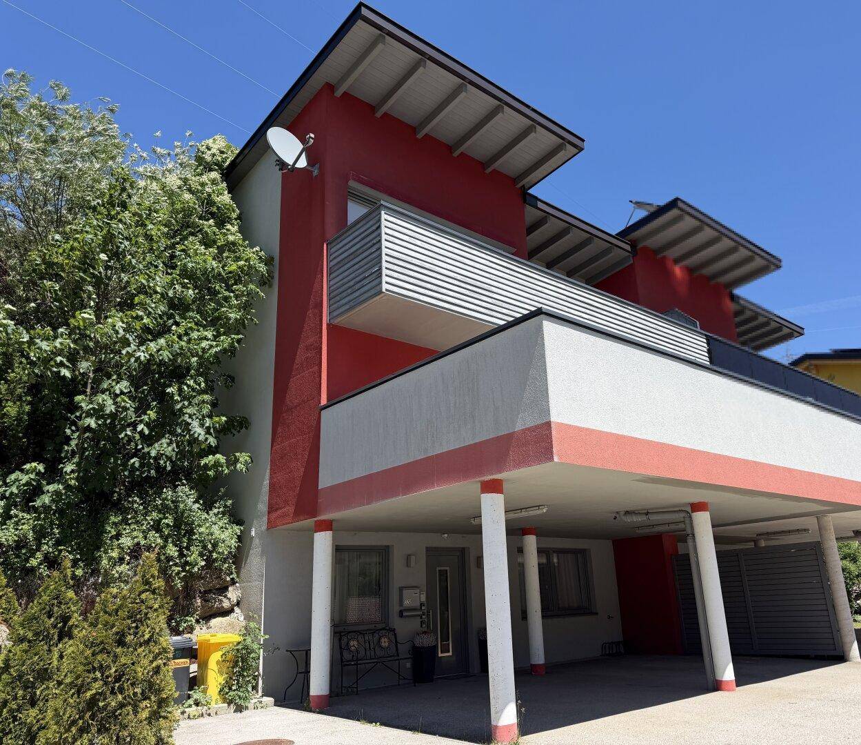 Moderne Gebäudefassade mit roten und grauen Elementen, Balkonen und einem überdachten Carport.
