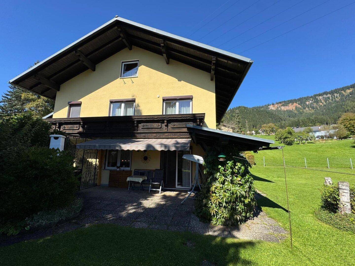 Traditionelles Haus mit gelber Fassade, überdachter Terrasse und Garten, umgeben von grünen Wiesen und Bergen.