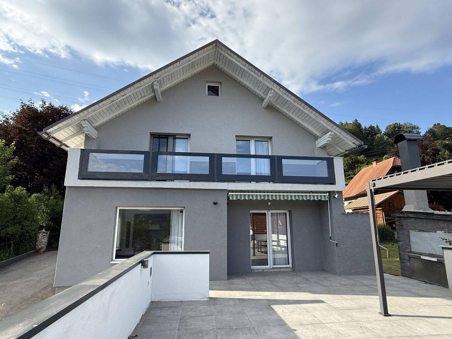 Modernes Einfamilienhaus mit großer gefliester Terrasse, Pergola und Balkon, umgeben von viel Grün.
