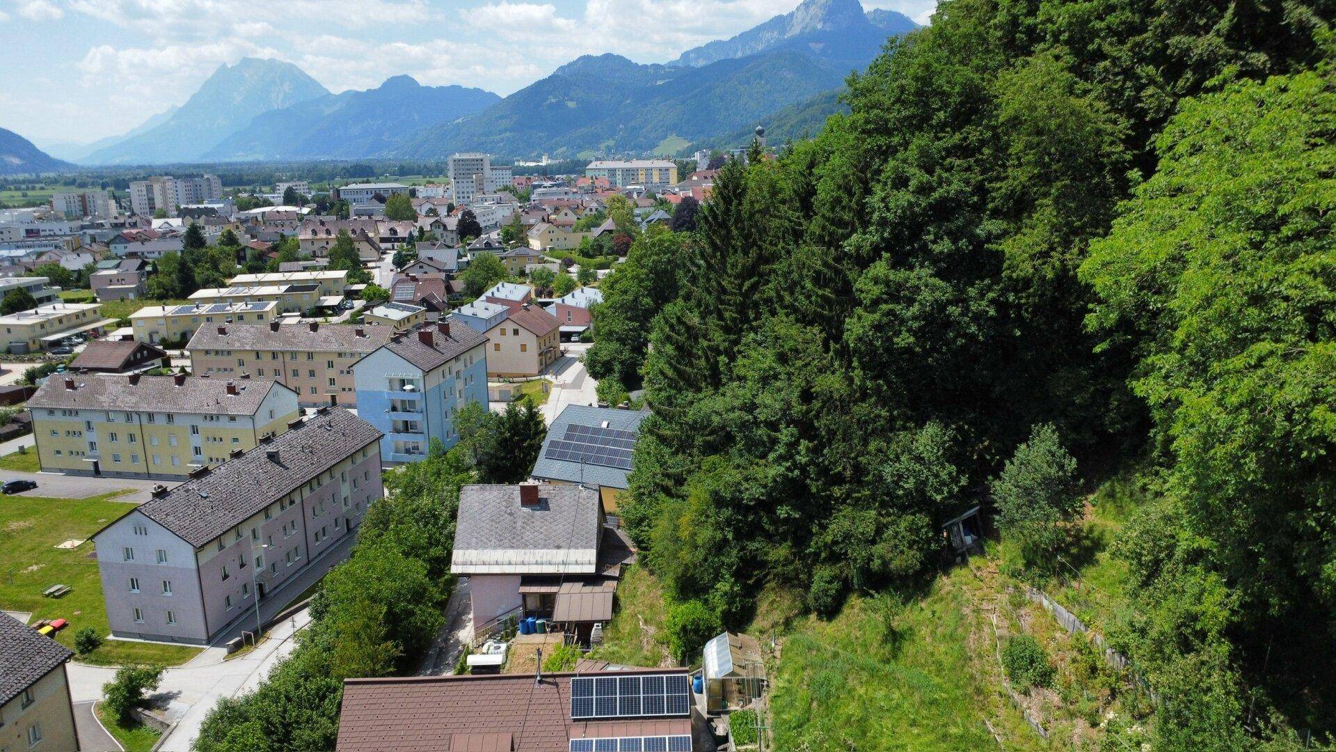 Weitläufige Luftaufnahme der Siedlung mit Häusern, Bergen und einer Stadt im Hintergrund unter blauem Himmel.