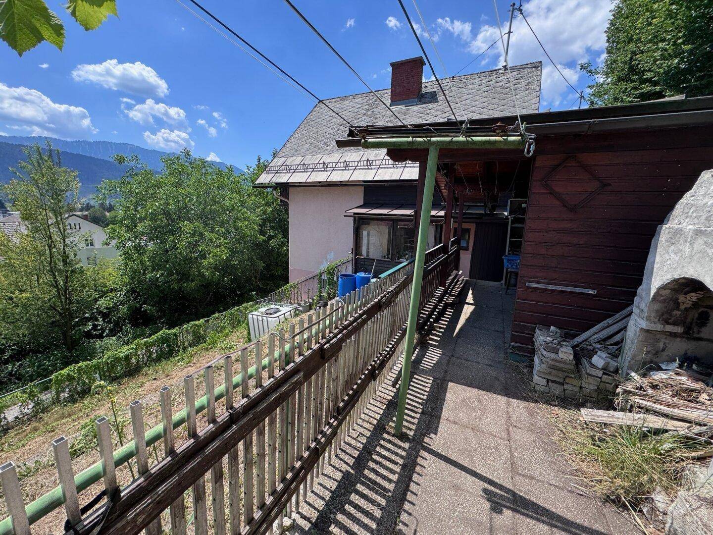 Außenbereich mit gepflastertem Weg entlang des Hauses und einem Holzschuppen im Hintergrund.