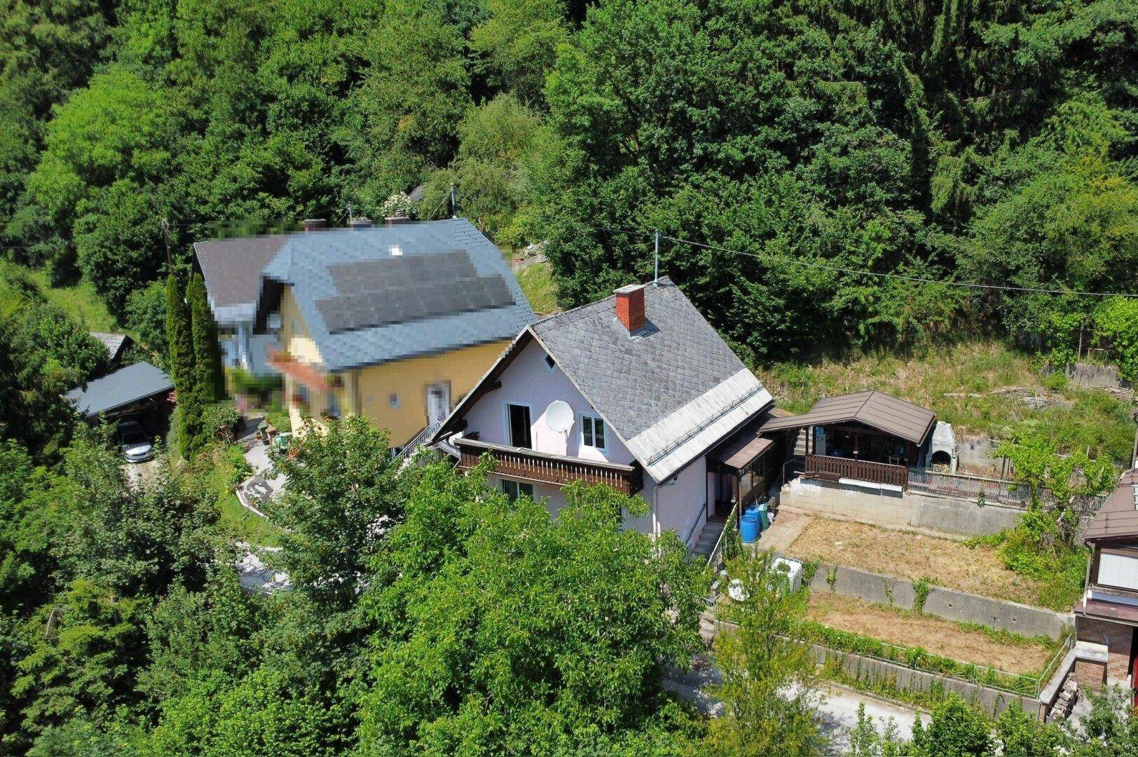 Luftaufnahme des Hauses mit Nachbargebäuden und viel Grün in einer bergigen Umgebung.