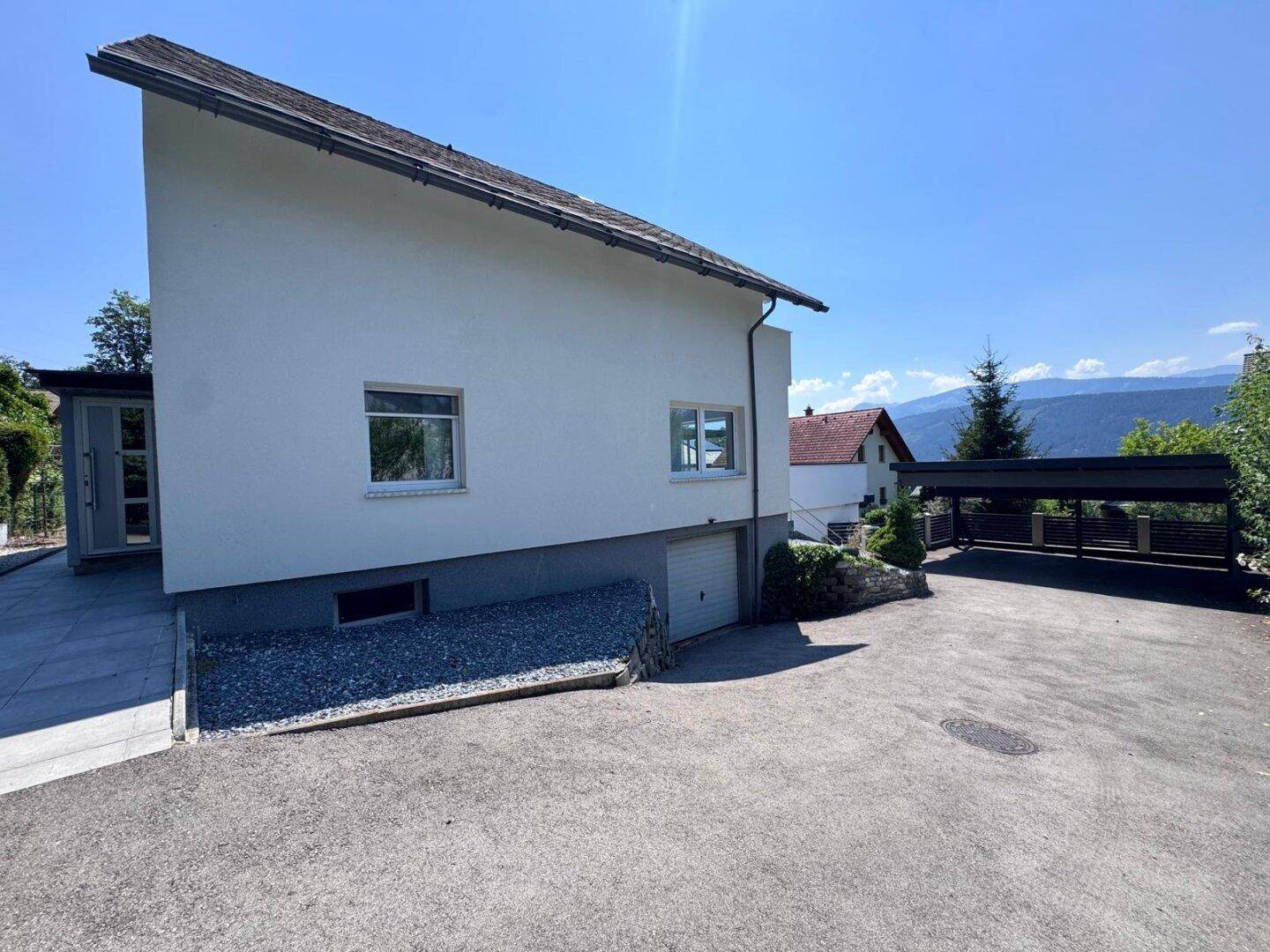 Seitenansicht des Hauses mit Carport, Zufahrt und Blick auf die umliegende Berglandschaft.