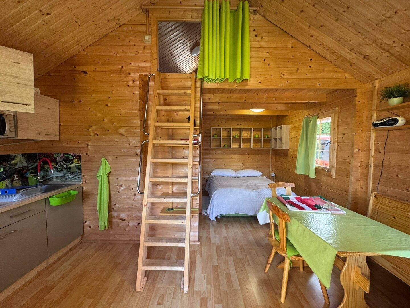 Offener Wohnbereich in einer Holzhütte mit Küchenzeile, Schlafbereich und Leiter zum Dachboden.