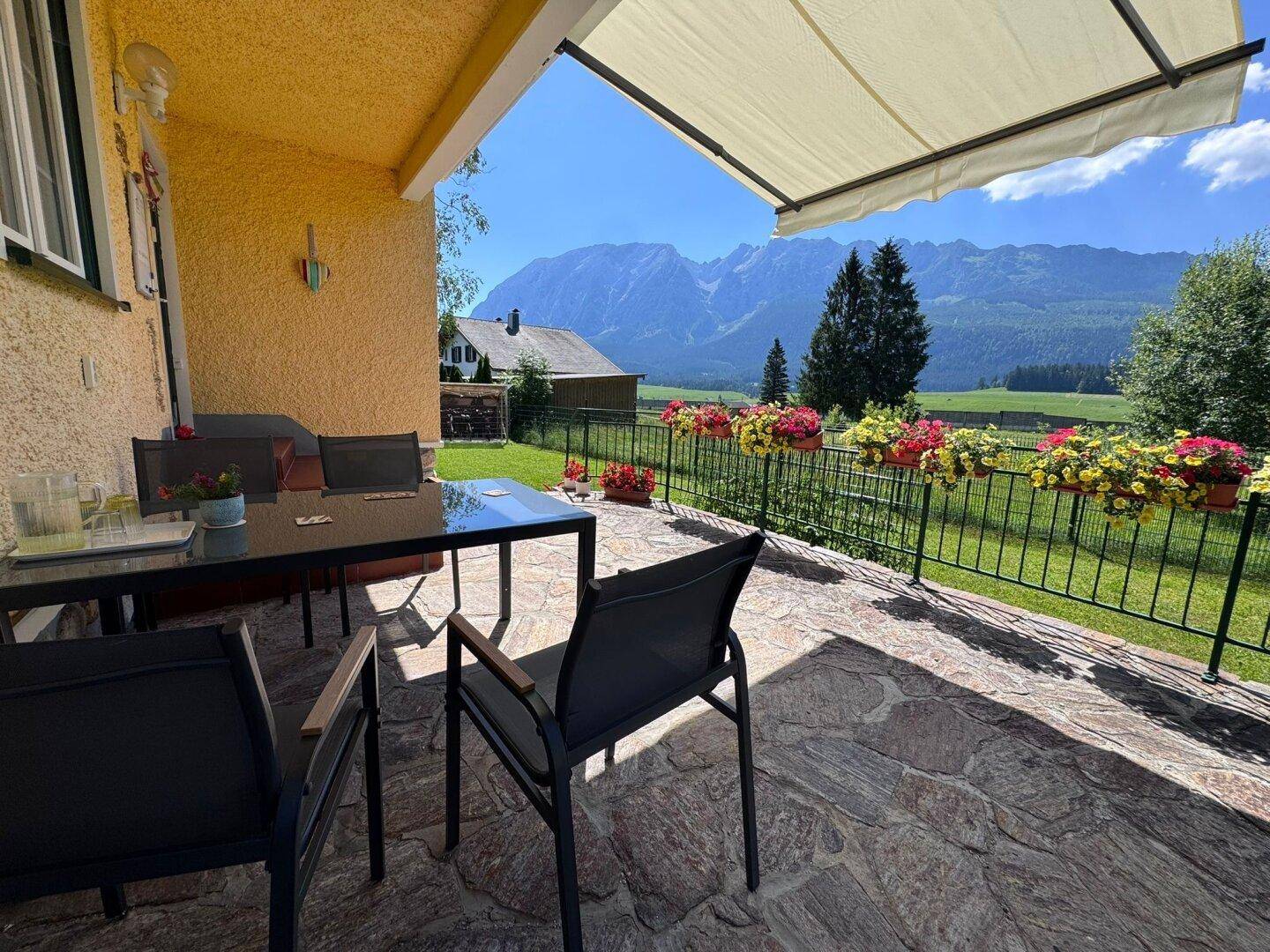 Gemütliche Terrasse mit Gartenmöbeln und Markise, umgeben von Blumen und Bergblick.