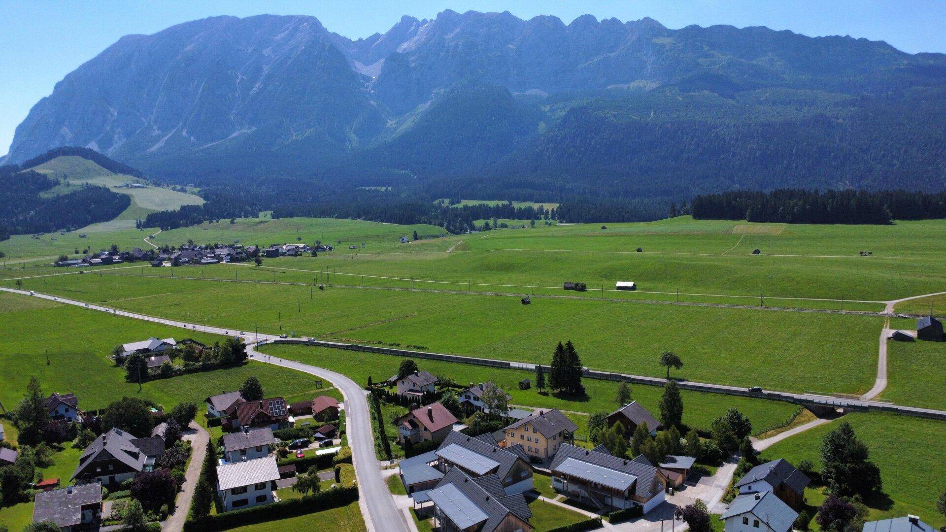 Panoramablick auf ein Dorf mit umliegenden Feldern und majestätischen Bergen im Hintergrund.