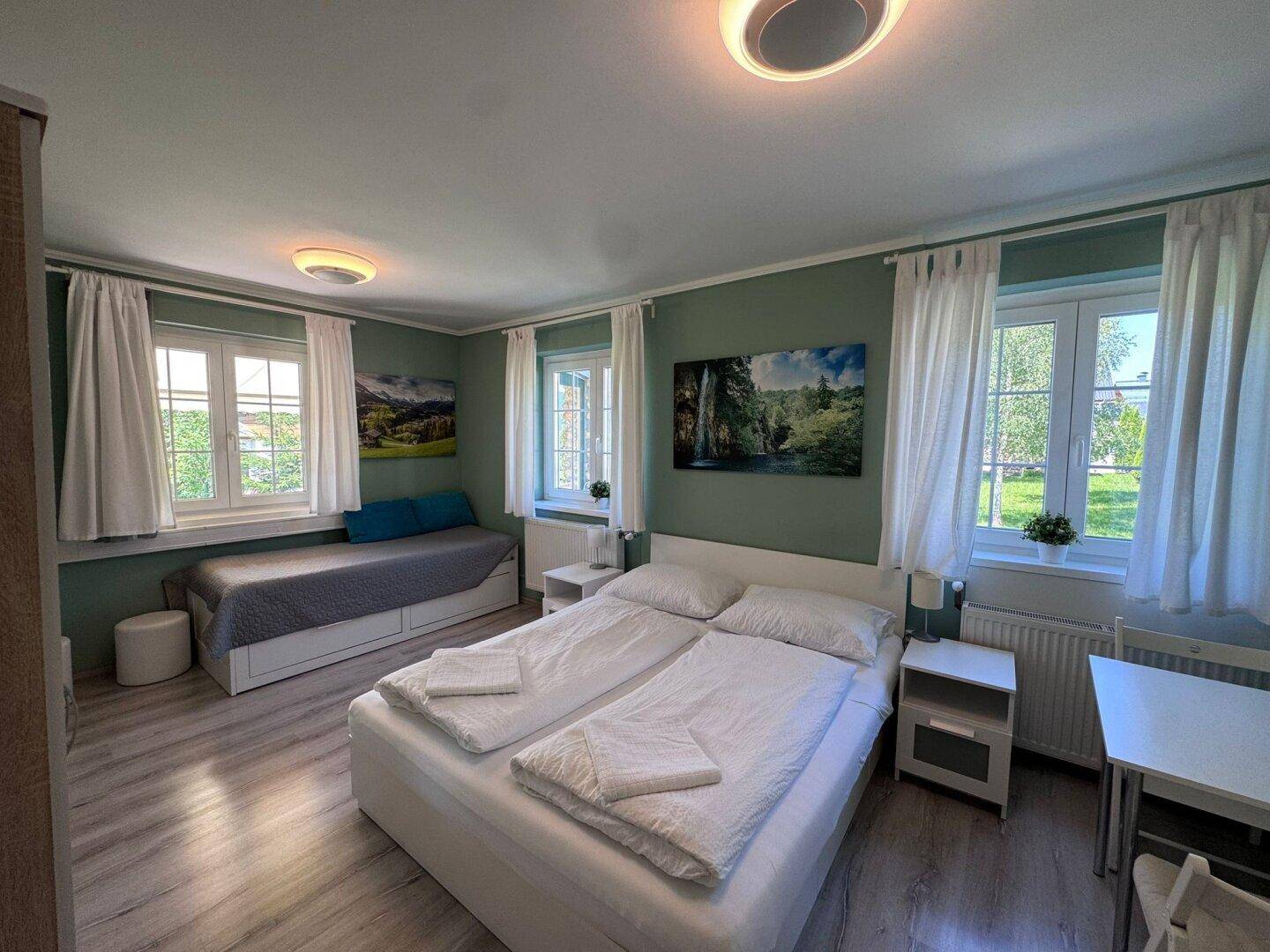 Helles Schlafzimmer mit Doppelbett, Tagesbett und grünen Wänden, bietet Blick ins Grüne.