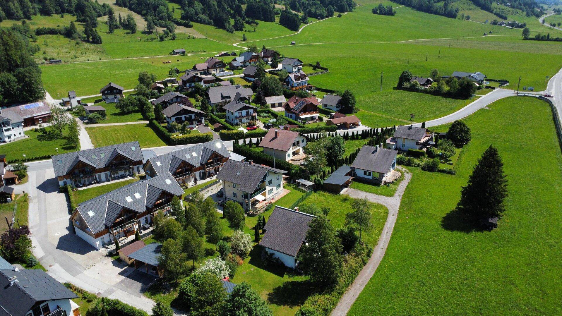 Panoramablick auf eine ländliche Wohnsiedlung mit Häusern und grünen Wiesen unter blauem Himmel.
