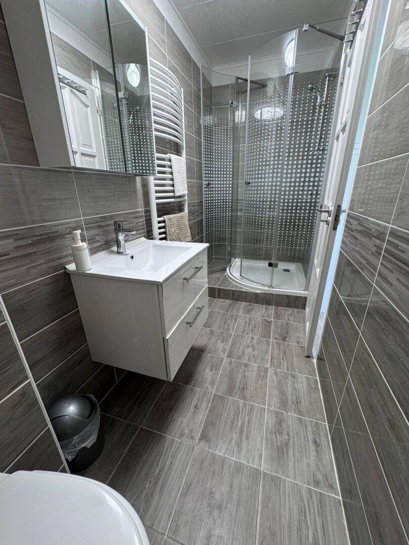 Modernes Badezimmer mit grauen Fliesen, Duschkabine, schwebendem Waschtisch und Handtuchheizkörper.